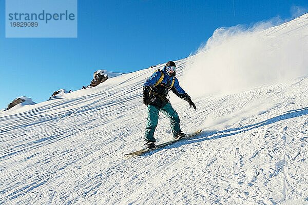 Ein Freeboard Snowboarder mit Skimaske und Rucksack fährt über die schneebedeckte Piste und hinterlässt einen Pulverschnee vor dem blaün Himmel