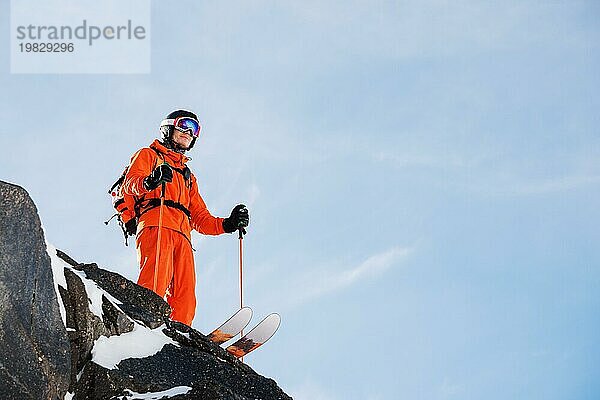 Ein professioneller Skisportler steht an einem sonnigen Tag am Rande eines hohen Felsens vor dem blaün Himmel. Kopieren Sie den Raum. Das Konzept des Skifahrens