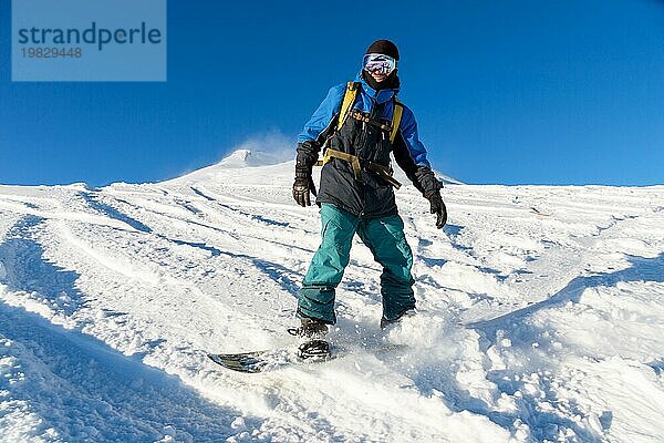 Ein Freeboard Snowboarder mit Skimaske und Rucksack fährt über die schneebedeckte Piste und hinterlässt einen Pulverschnee vor dem blaün Himmel