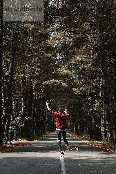 Ein junger Hipster mit Mütze und kariertem Hemd vollführt einen Trick  indem er auf zwei Rädern seines Longboards auf einer Landstraße in einem herbstlichen Wald balanciert. Das Konzept des Reisens mit leichtem Gepäck und Extremsportarten in der Natur