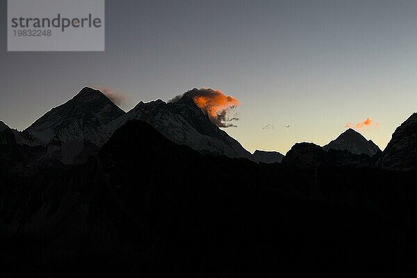Blick in Richtung Osten bei Tagesanbruch vom Gokyo Ri  dem beliebten Trekking Gipfel und Aussichtspunkt in der Everest Region. Die Silhouetten von drei Achttausendern sind zu sehen  die von der aufgehenden Sonne angestrahlt werden: Der Mount Everest befindet sich unter der orangefarbenen Wolke auf der linken Seite  dann folgt der Lhotse  der vierthöchste Berg der Erde. Der Makalu mit seiner pyramidenartigen Form ist weiter rechts zu sehen. Gokyo Trek  Khumbu  Everest Region  Himalaya. Der Sagarmatha Nationalpark  ein UNESCO Welterbe. Solukhumbu  Nepal  Asien
