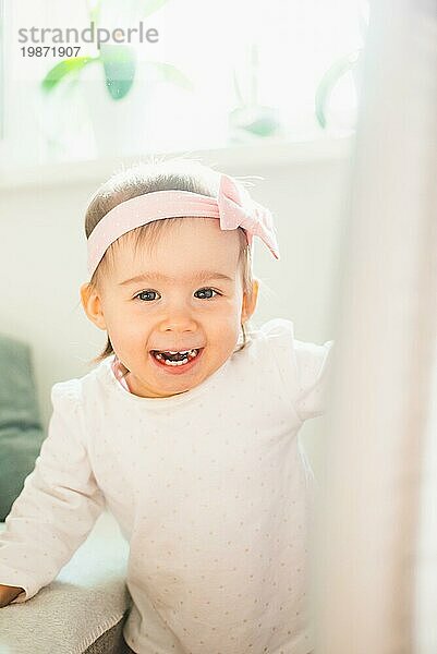Kaukasische Baby Mädchen in hellen Raum  natürliches Licht. Porträt des Kindes mit rosa Stirnband Bogen. Schaut in die Kamera und lacht. Vertikale Fotografie