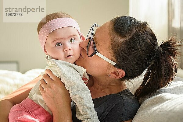 Mutter hält und küsst schönes Baby Mädchen in hellen sonnigen Raum. Mutterschaft Hintergrund