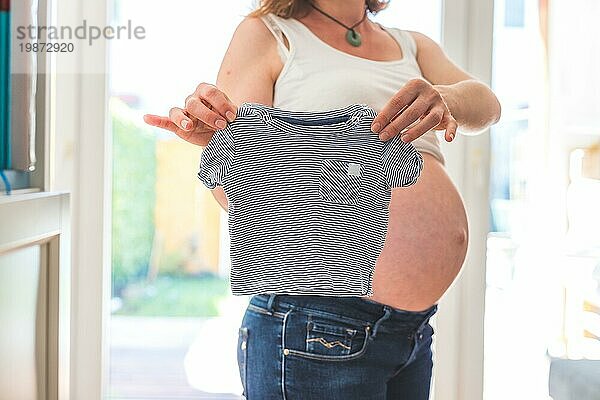 Glückliche kaukasische schwangere Mutter mit kleinem Baby Shirt