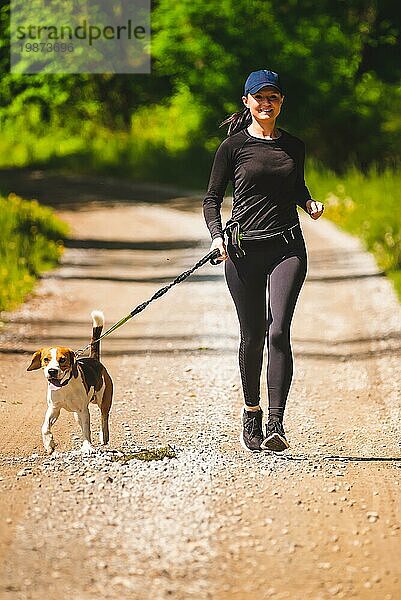 Sportliches Mädchen läuft mit einem Hund (Beagle) an einem sonnigen Frühlingstag auf einem Waldweg. Raum in der Natur kopieren