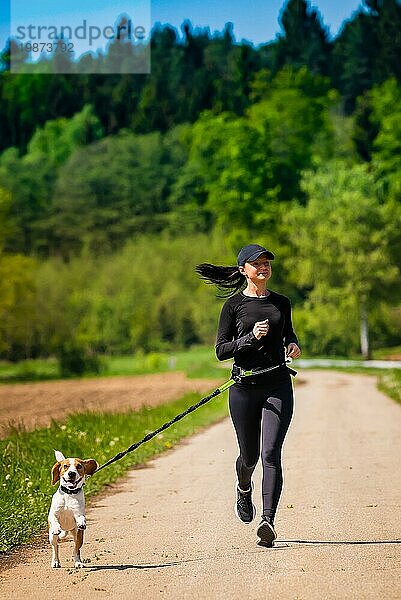 Sportliches Mädchen läuft mit einem Hund (Beagle) an der Leine im Frühling  sonniger Tag auf der Landstraße zum Wald. Kopieren Raum in der Natur