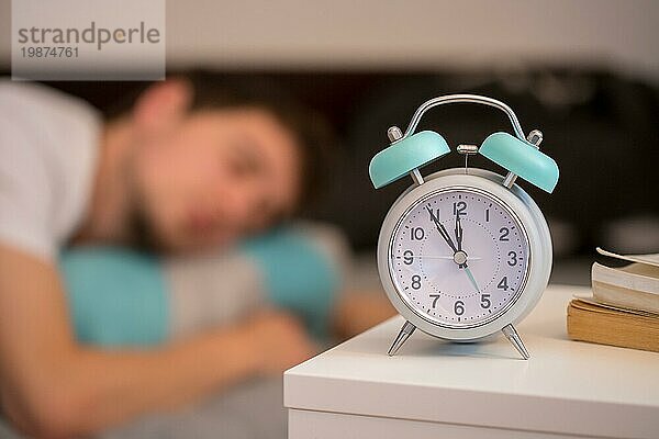 Weißer Wecker auf dem Nachttisch. Junger Mann schläft im Hintergrund