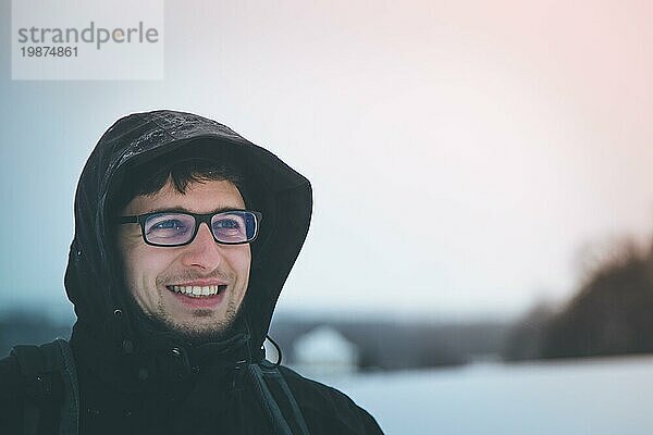 Junger lächelnder Mann in schwarzer Jacke mit Kapuze  im Winter draußen