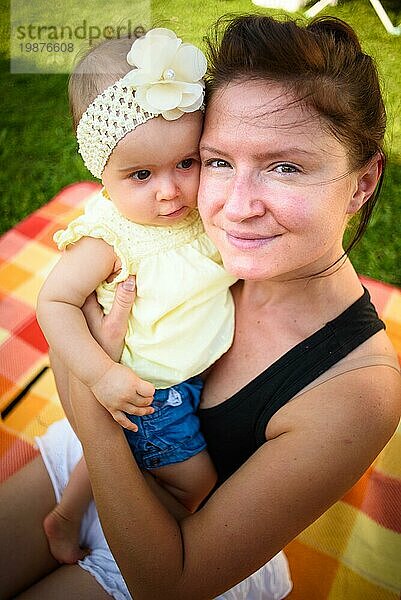 Portrait junge Mutter hält niedlichen Baby Mädchen in gelben Band und Kleid. Sonniger Sommertag im Freien. Mutterschaft Konzept