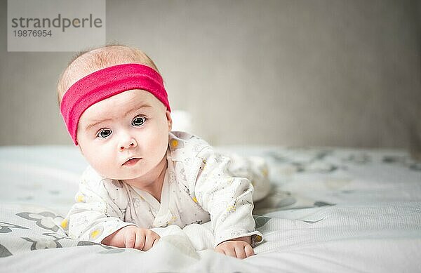 Adorable 6 Monate alt Baby Mädchen Kleinkind auf einem Bett auf dem Bauch mit dem Kopf nach oben in die Kamera mit ihren großen Augen. Natürliches Schlafzimmer Licht