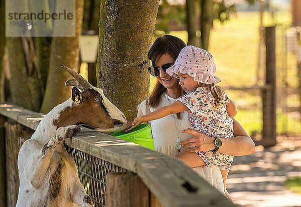 Glückliches Kind  das von der Mutter gehalten wird  die eine Ziege auf dem Bauernhof aus der Hand füttert. Kind Entwicklung Konzept
