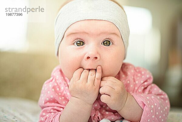 Nettes 6 Monate altes Baby Mädchen auf einem Bett auf dem Bauch mit dem Kopf nach oben und die Finger in den Mund  Blick in Richtung Kamera mit ihren großen Augen