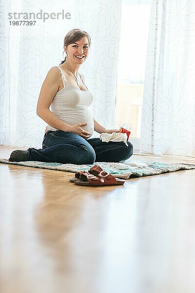 Glückliche kaukasische schwangere Mutter sitzt auf dem Boden  berührt ihren Bauch  Blue Jeans