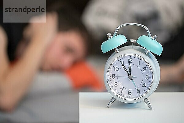 Weißer Wecker auf dem Nachttisch. Junge versucht  im Hintergrund zu schlafen  Schlaflosigkeit