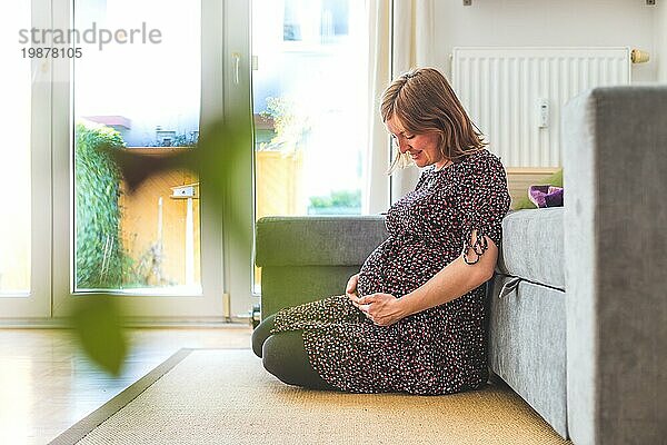 Schwangere kaukasische glückliche Mutter sitzt auf dem Boden  berührt ihren Bauch  blumiges Kleid