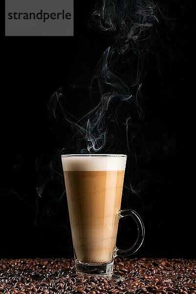 Heißer dampfender Milchkaffee mit schaumiger Milch in einem hohen Glas  auf Kaffeebohnen vor schwarzem Hintergrund. Ansicht von oben