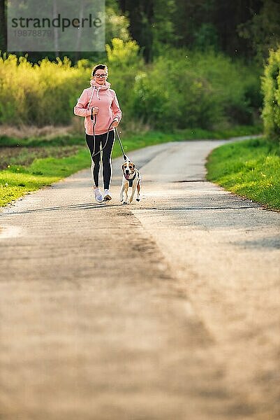 Sportliches Mädchen läuft mit einem Hund (Beagle) an der Leine im Frühling  sonniger Tag auf der Landstraße zum Wald. Kopieren Raum in der Natur