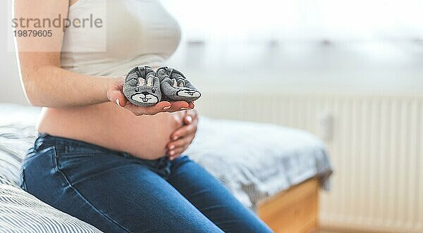 Caucasian schwangere Mutter hält Baby Schuhe in ihren Händen  nackten Bauch in der unscharfen Hintergrund