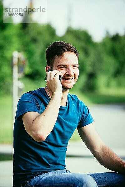 Hübscher junger Mann sitzt im Freien und spricht mit jemandem am Telefon