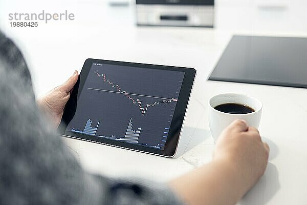 Frau schaut auf ein digitales Tablet mit Börsendiagramm und hält eine Tasse Kaffee in einer modernen Küche. Trading zu Hause Konzept