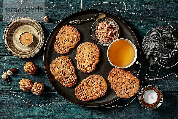 Halloween Ingwerplätzchen in Form von Totenköpfen  hausgemachte Dia de los muertos Kekse  von oben aufgenommen mit Tee und Kerzen auf dunklem Holzhintergrund