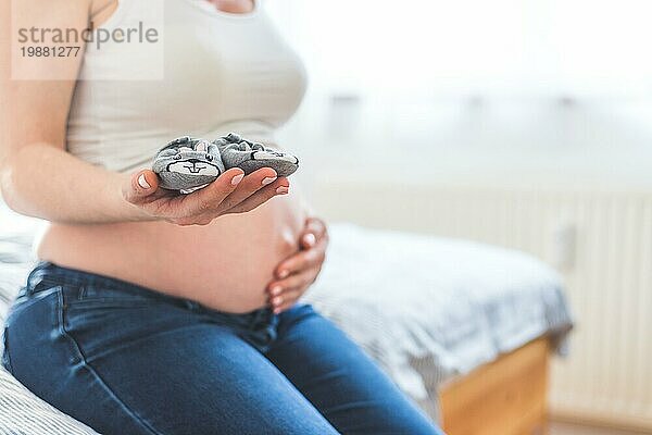 Caucasian schwangere Mutter hält Baby Schuhe in ihren Händen  nackten Bauch in der unscharfen Hintergrund