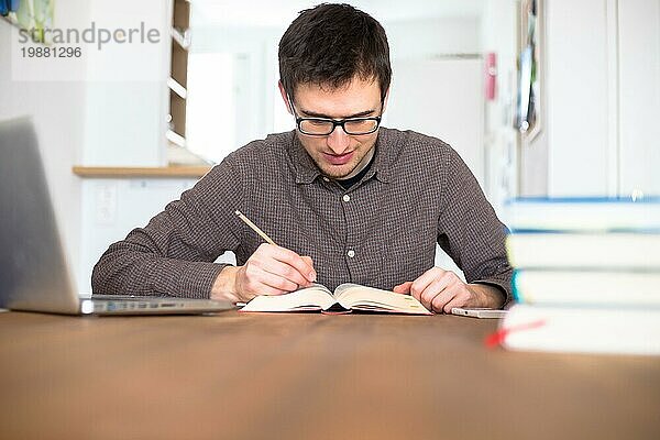 Männlicher glücklicher Student  der zu Hause recherchiert und lernt  Stapel von Büchern unscharf