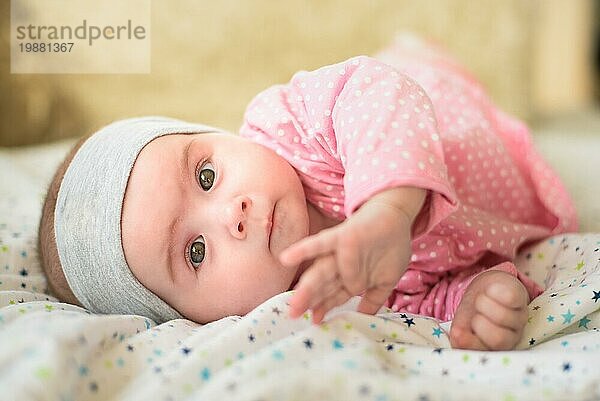 Cute 6 Monate alt Baby Mädchen Kleinkind auf einem Bett auf dem Bauch mit dem Kopf nach oben in die Kamera mit ihren großen Augen. Natürliches Schlafzimmer Licht