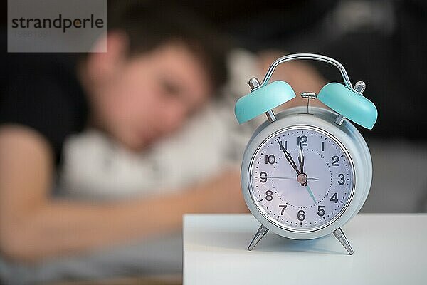 Weißer Wecker auf dem Nachttisch. Junger Mann schläft im Hintergrund