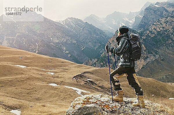 Ein Mann mit Bart und Sonnenbrille in einer Membranjacke  Hut  mit einem Rucksack und Stöcken für Nordic Walking  ein Reisender  der in der freien Natur steht und auf die Berge schaut