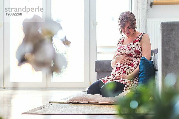 Schwangere kaukasische glückliche Mutter sitzt auf dem Boden  berührt ihren Bauch  blumiges Kleid