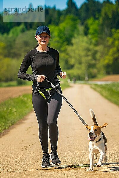 Sportliches Mädchen läuft mit einem Hund (Beagle) an einem sonnigen Frühlingstag auf einem Waldweg. Raum in der Natur kopieren