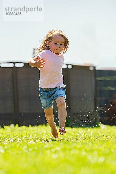 Fröhliches Kind läuft durch naßen Rasen und spritzt mit Wasser. Kindheit Konzept