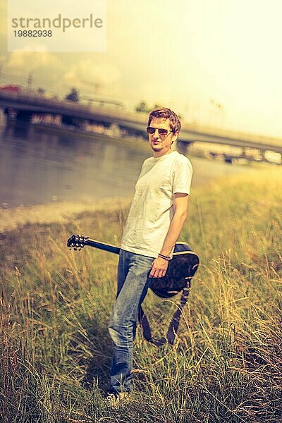Junger Mann sitzt an einem Fluss und spielt auf seiner Westerngitarre