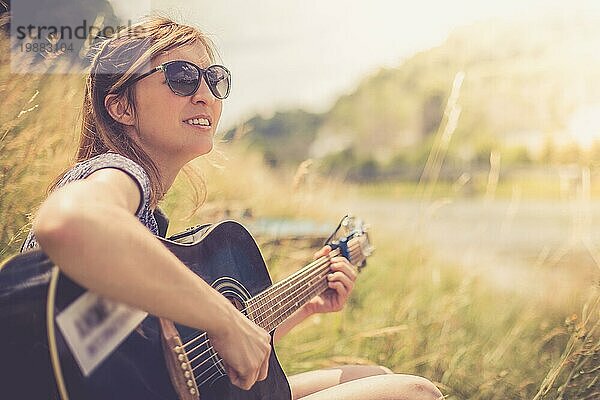 Schönes junges Mädchen sitzt an einem Fluss und spielt ihre Westerngitarre