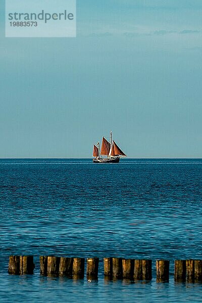 Landschaftliches Meeresbild mit einem Segelboot mit braunen Segeln am Horizont und einer Buhne im Vordergrund