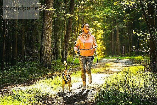 Junge Frau und Hund laufen zusammen im sonnigen Wald. Fröhliche Frau  die mit ihrem Haustier im Freien trainiert. Aktive Menschen Konzept