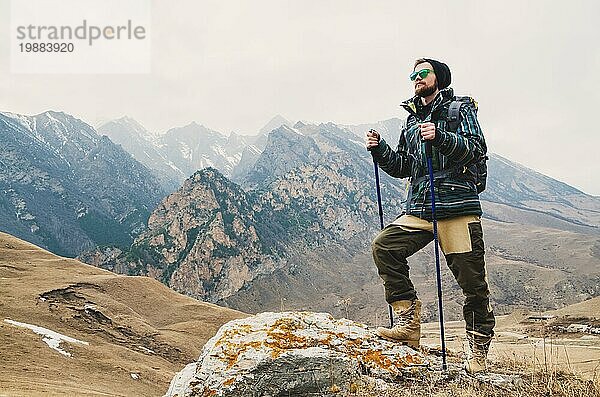 Ein Mann mit Bart und Sonnenbrille in einer Membranjacke  Hut  mit einem Rucksack und Stöcken für Nordic Walking  ein Reisender  der in der freien Natur steht und auf die Berge schaut