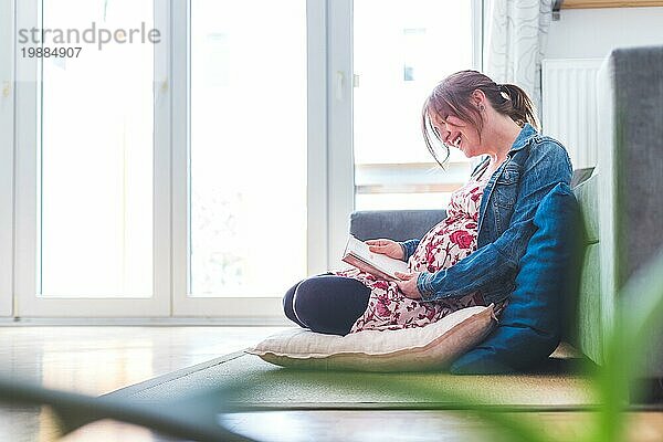 Schwangere kaukasische glückliche Mutter sitzt auf dem Boden  berührt ihren Bauch  blumiges Kleid