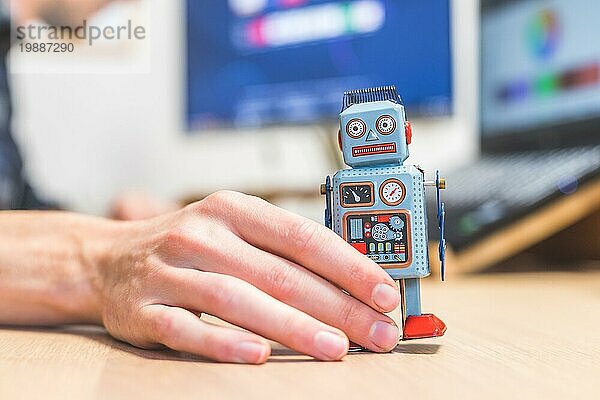 Spielzeugroboter auf einem Schreibtisch  im Hintergrund arbeitet ein Mann am Computer. Symbol für künstliche Intelligenz  Chatbots oder soziale Bots und Algorithmen