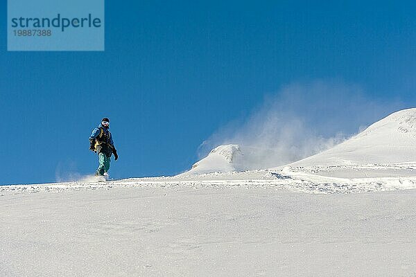 Ein Freeboard Snowboarder mit Skimaske und Rucksack fährt über die schneebedeckte Piste und hinterlässt einen Pulverschnee vor dem blaün Himmel