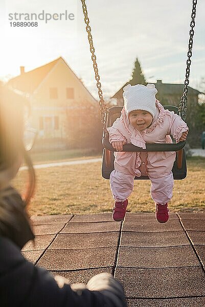 Adorable Baby Mädchen mit großen schönen Augen und eine Mütze mit Spaß auf einer Schaukel Fahrt auf einem Spielplatz in einem sonnigen Park