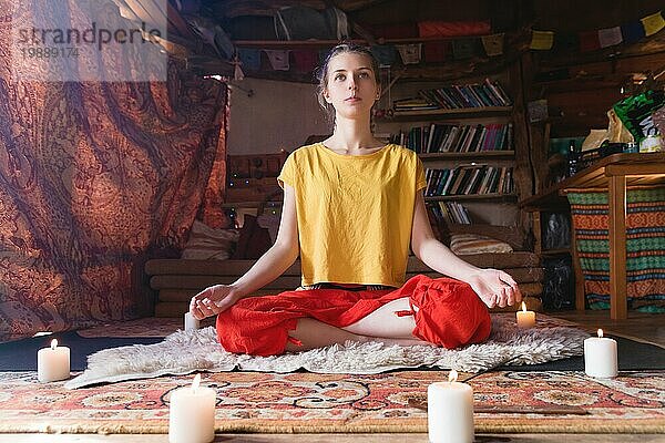 Porträt eines jungen Mädchens in heller Kleidung  das in einem von Kerzen umgebenen Bastelraum meditiert. Newage Richtung und spirituelle Entwicklung