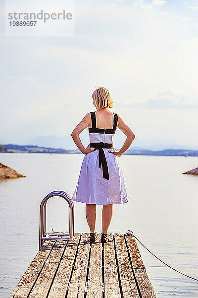 Junges Mädchen mit weißem Kleid steht auf einem Steg und genießt die Aussicht auf den See