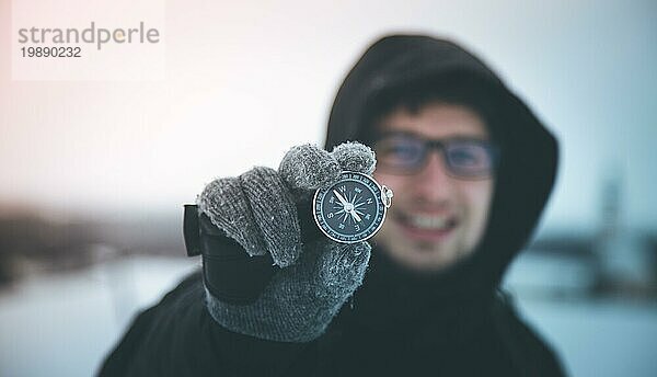 Ein junger lächelnder Mann hält einen Kompass hoch. Winterzeit  Handschuhe und Kapuze