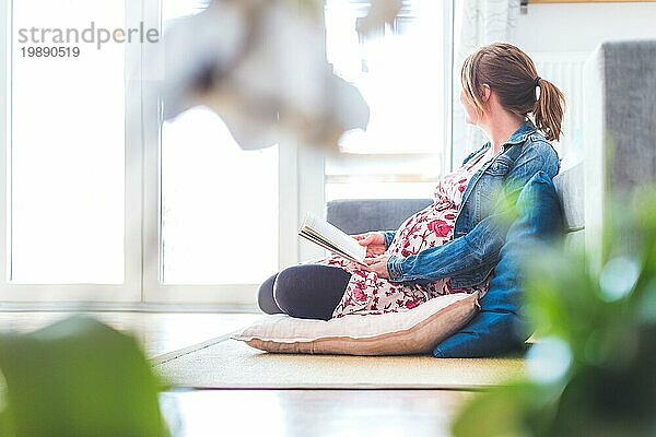 Schwangere kaukasische glückliche Mutter sitzt auf dem Boden und liest ein Buch