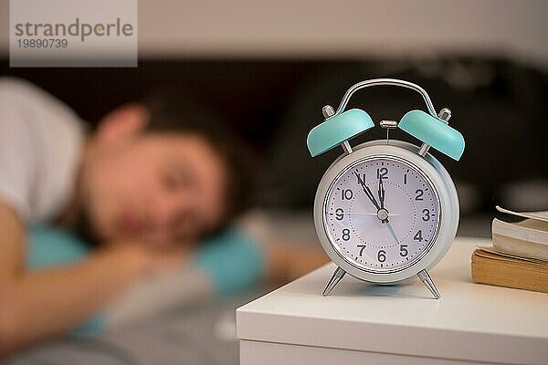 Weißer Wecker auf dem Nachttisch. Junger Mann schläft im Hintergrund