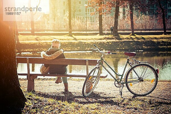 Rückenansicht einer jungen Frau mit Fahrrad  die die Sonne auf einer Parkbank genießt  Frühling