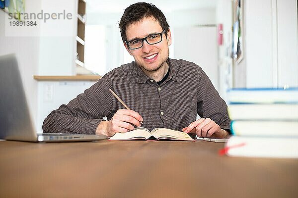 Männlicher glücklicher Student  der zu Hause recherchiert und lernt  Stapel von Büchern unscharf