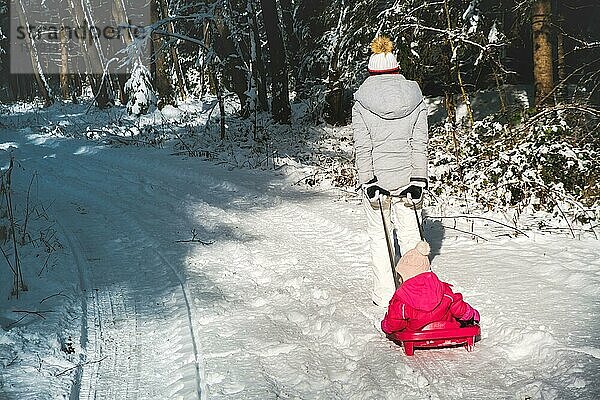 Mutter  die ihr Baby auf einem Schlitten durch den Winterwald zieht. Thema verschneiter Wald